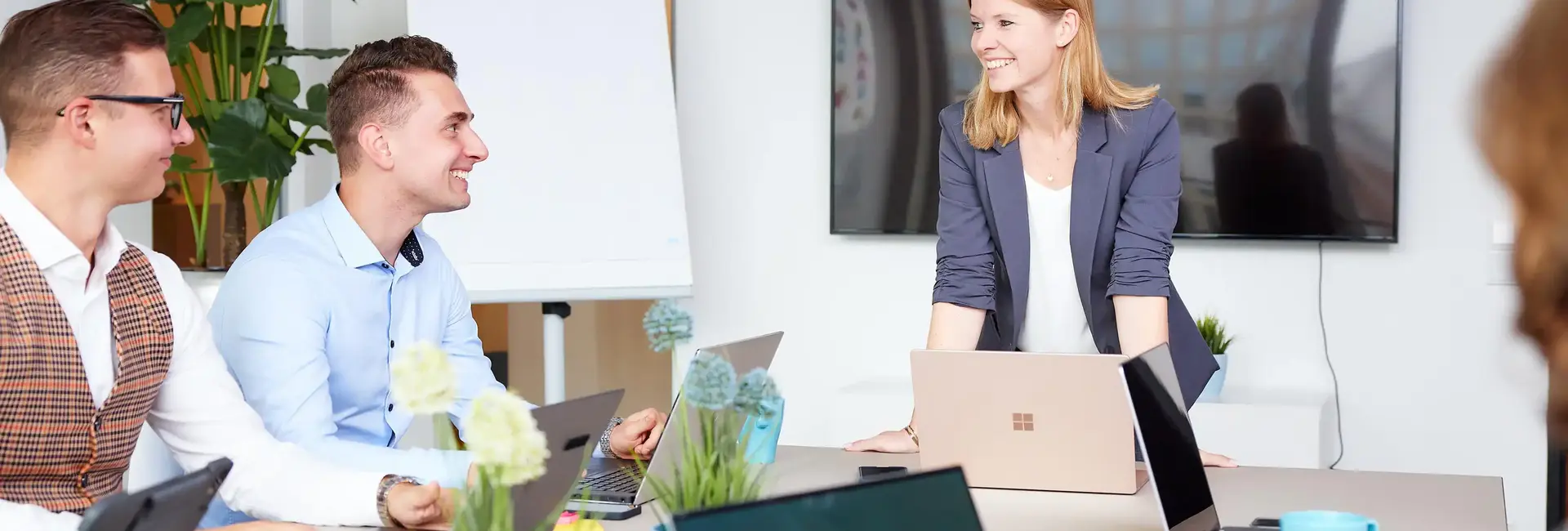 Eine Frau steht lächelnd an einem Tisch mit Laptops, während drei Personen ihr aufmerksam zuhören. Der Raum ist modern eingerichtet mit Pflanzen und Whiteboard.