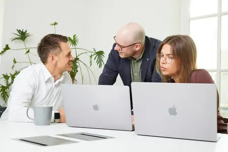 Drei Personen sitzen an einem Tisch mit Laptops und besprechen etwas. Eine Person steht und beugt sich vor, während die anderen aufmerksam zuhören.