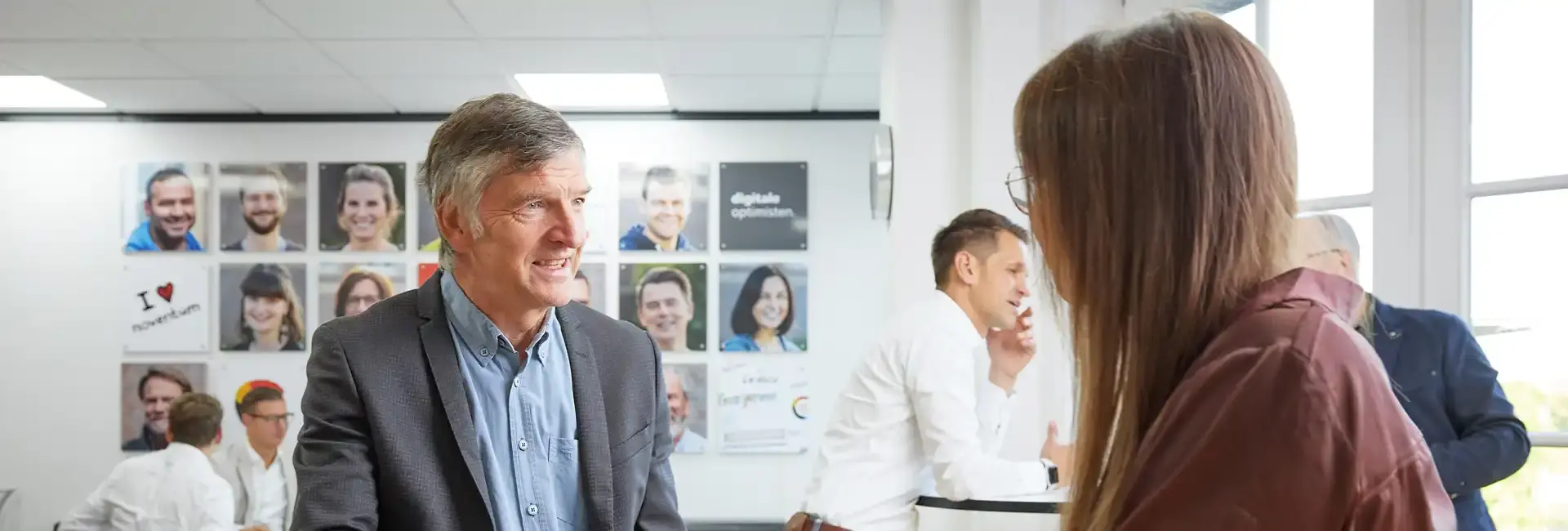 Ein Mann in einem Sakko spricht lächelnd mit einer Frau. Im Hintergrund sind Mitarbeitende in einem Büro mit einer Wand voller Porträtfotos zu sehen.