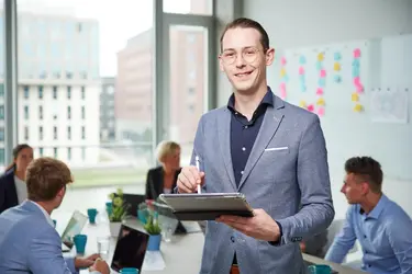 Ein junger Mann in Business-Outfit steht mit einem Tablet in der Hand und lächelt. Im Hintergrund arbeitet ein Team an einem Tisch mit Laptops und Notizen.