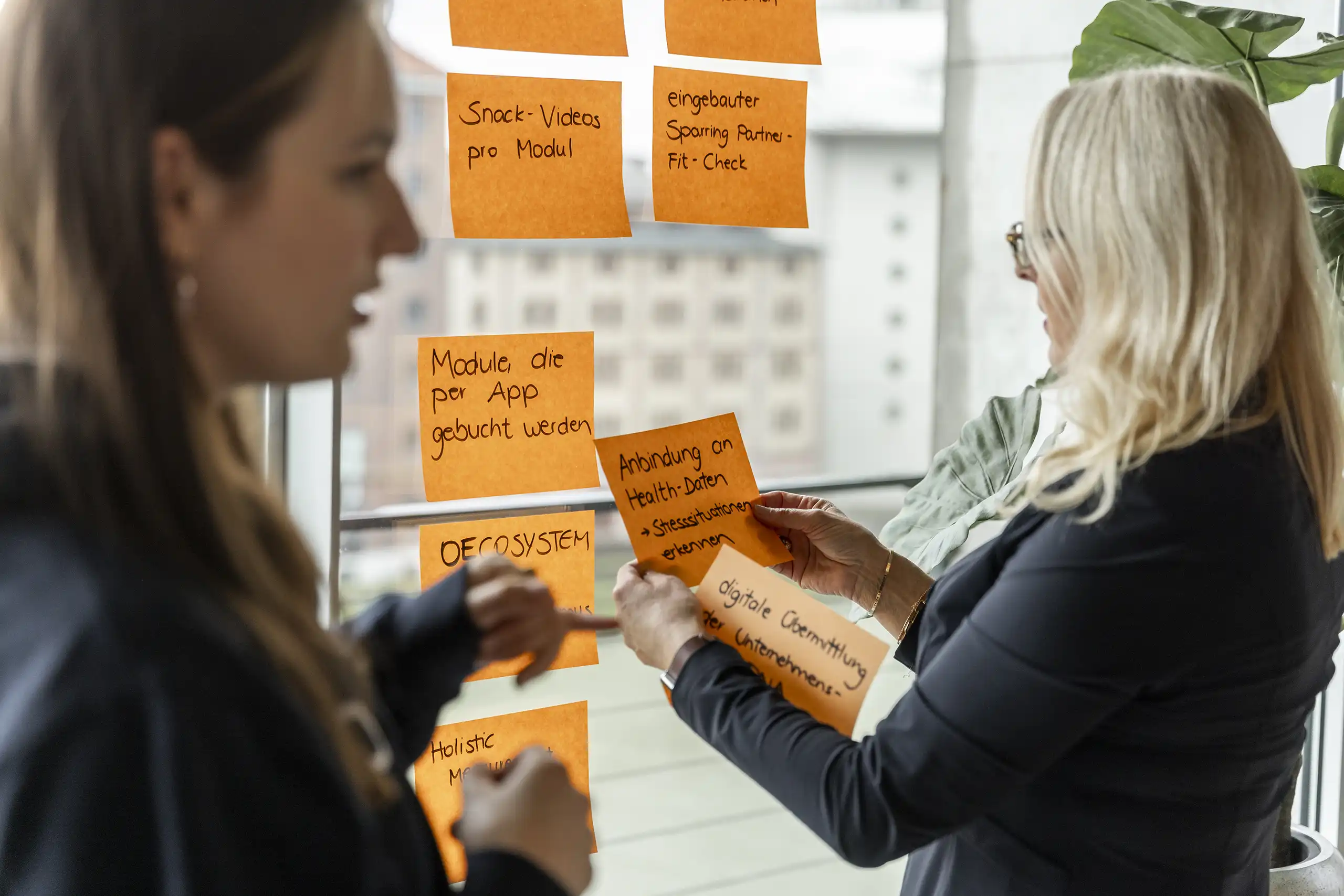 Zwei Frauen arbeiten an einer Glaswand mit orangefarbenen Notizzetteln. Eine hält Zettel in der Hand, während die andere gestikuliert und auf die Notizen zeigt