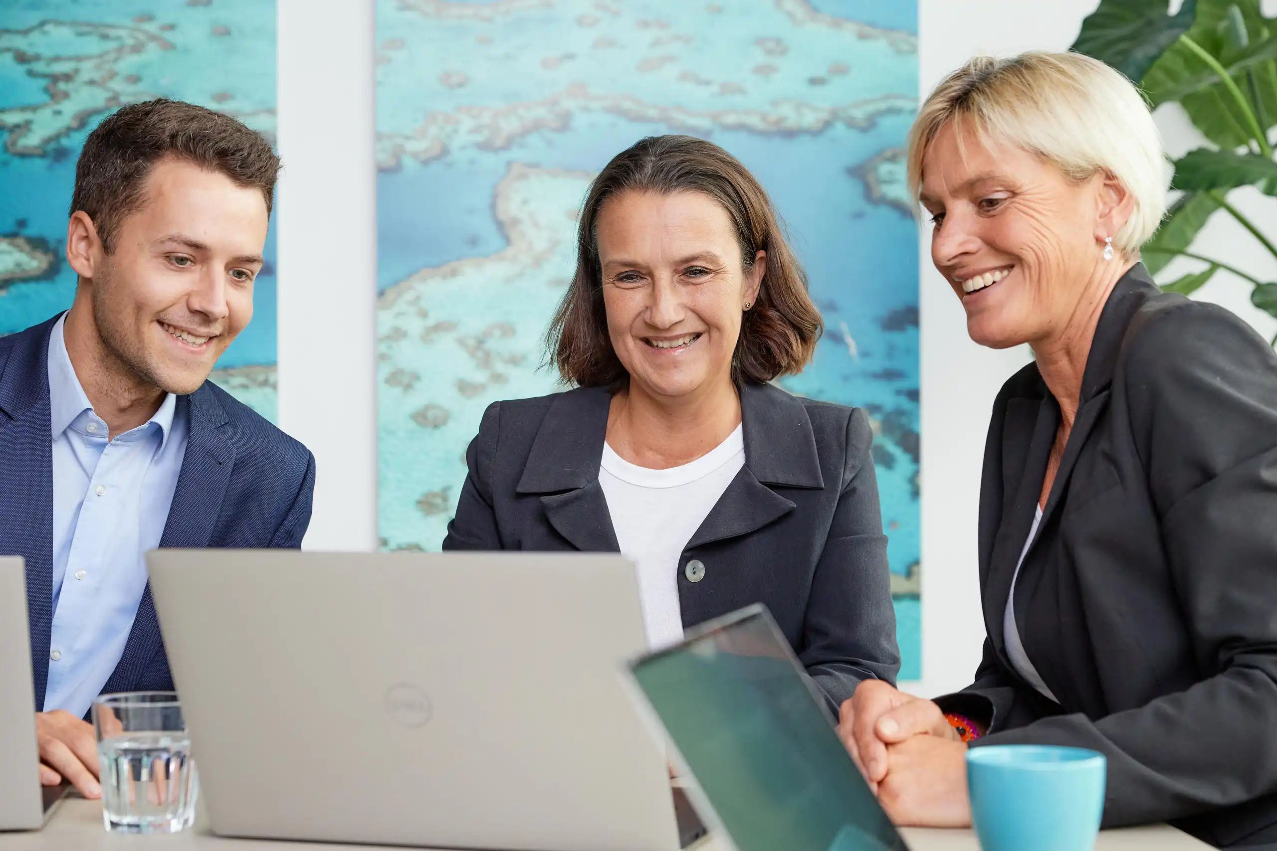 Drei Personen in Business-Kleidung sitzen gemeinsam an einem Tisch mit Laptops. Sie lachen und schauen auf einen Bildschirm, während sie sich austauschen.