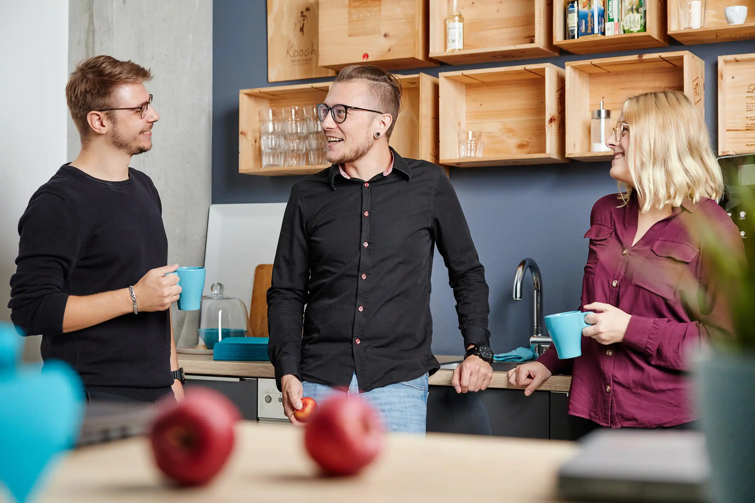 Drei Personen unterhalten sich entspannt in einer modernen Büroküche. Sie halten Tassen oder Obst in der Hand und lachen.