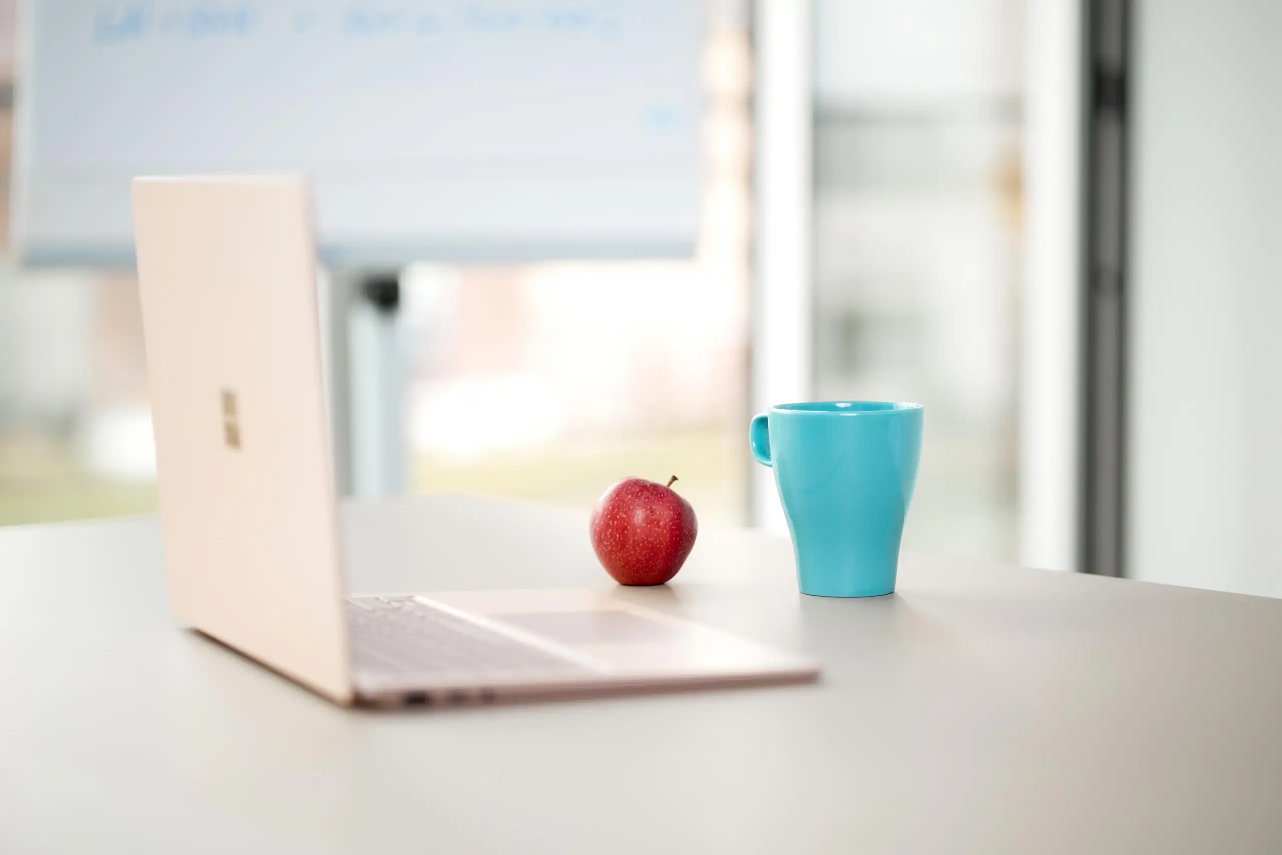 Ein aufgeschlagener Laptop, ein roter Apfel und eine blaue Tasse stehen auf einem Schreibtisch in einem modernen Büro. Im Hintergrund ist ein Whiteboard zu sehen.