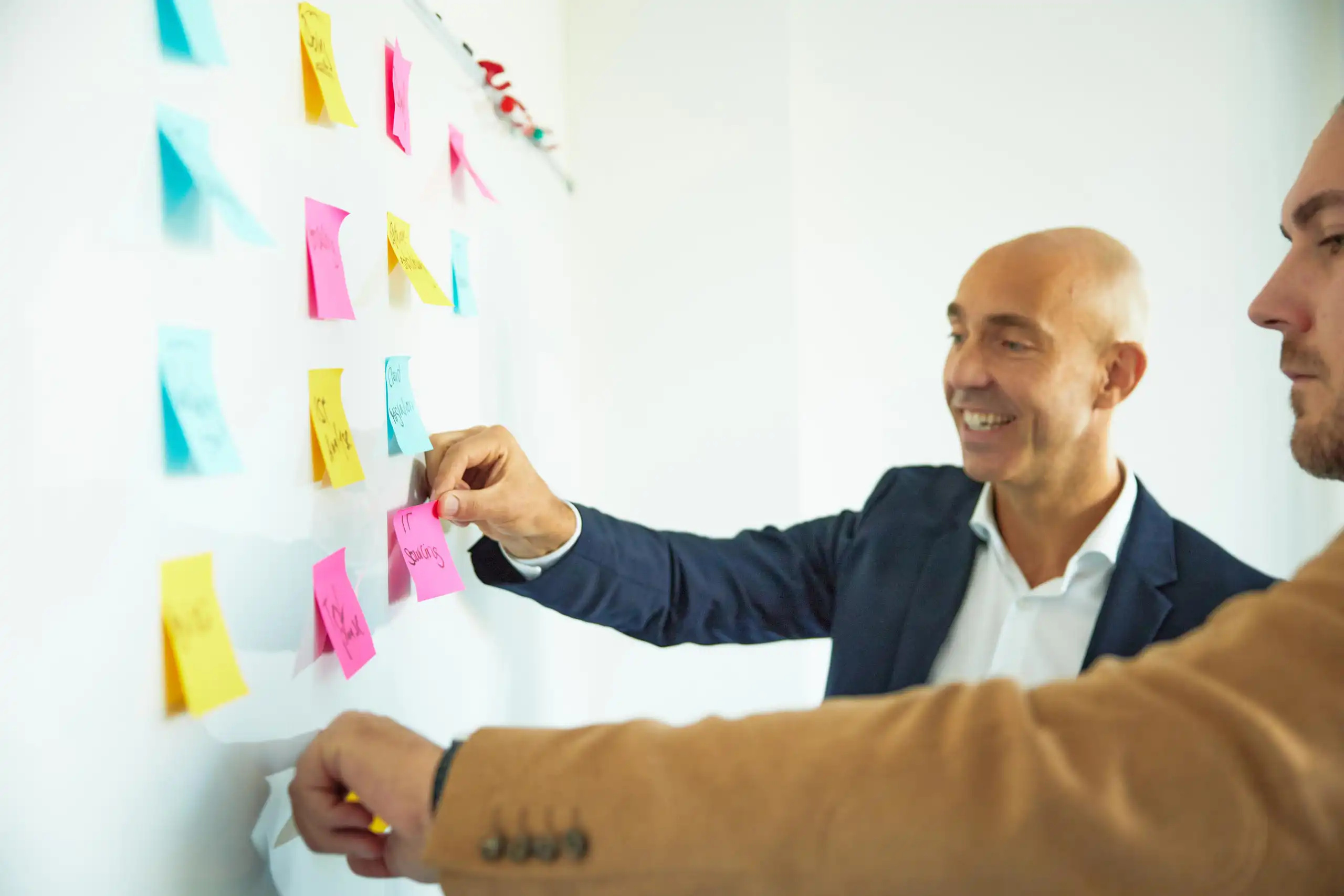 Zwei Männer in Business-Kleidung arbeiten lächelnd an einer Pinnwand mit bunten Post-Its.