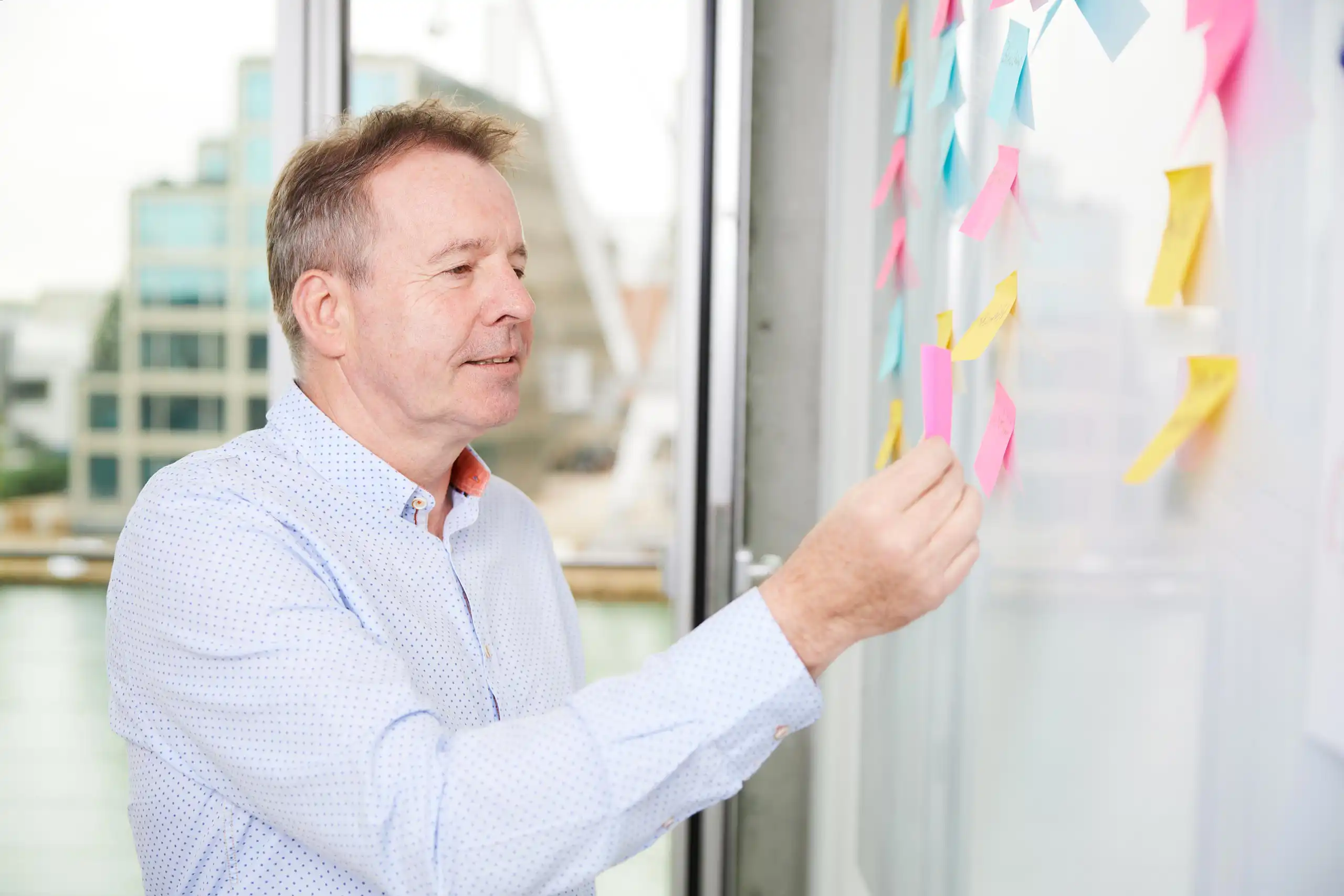 Ein Mann in einem hellen Hemd klebt eine bunte Haftnotiz an ein Whiteboard. Er wirkt konzentriert und arbeitet an einem Brainstorming in einem modernen Büro.