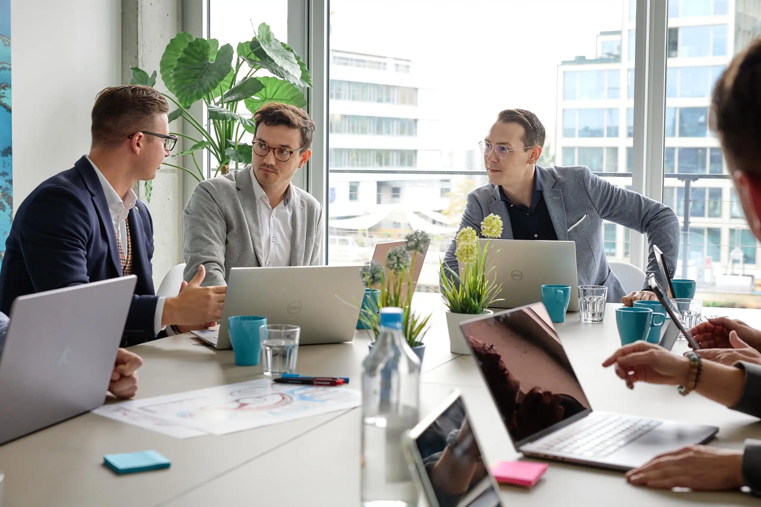 Mehrere Personen sitzen in einem modernen Büro an einem Tisch mit Laptops, Pflanzen und Notizen. Sie diskutieren konzentriert in einer Besprechungssituation.